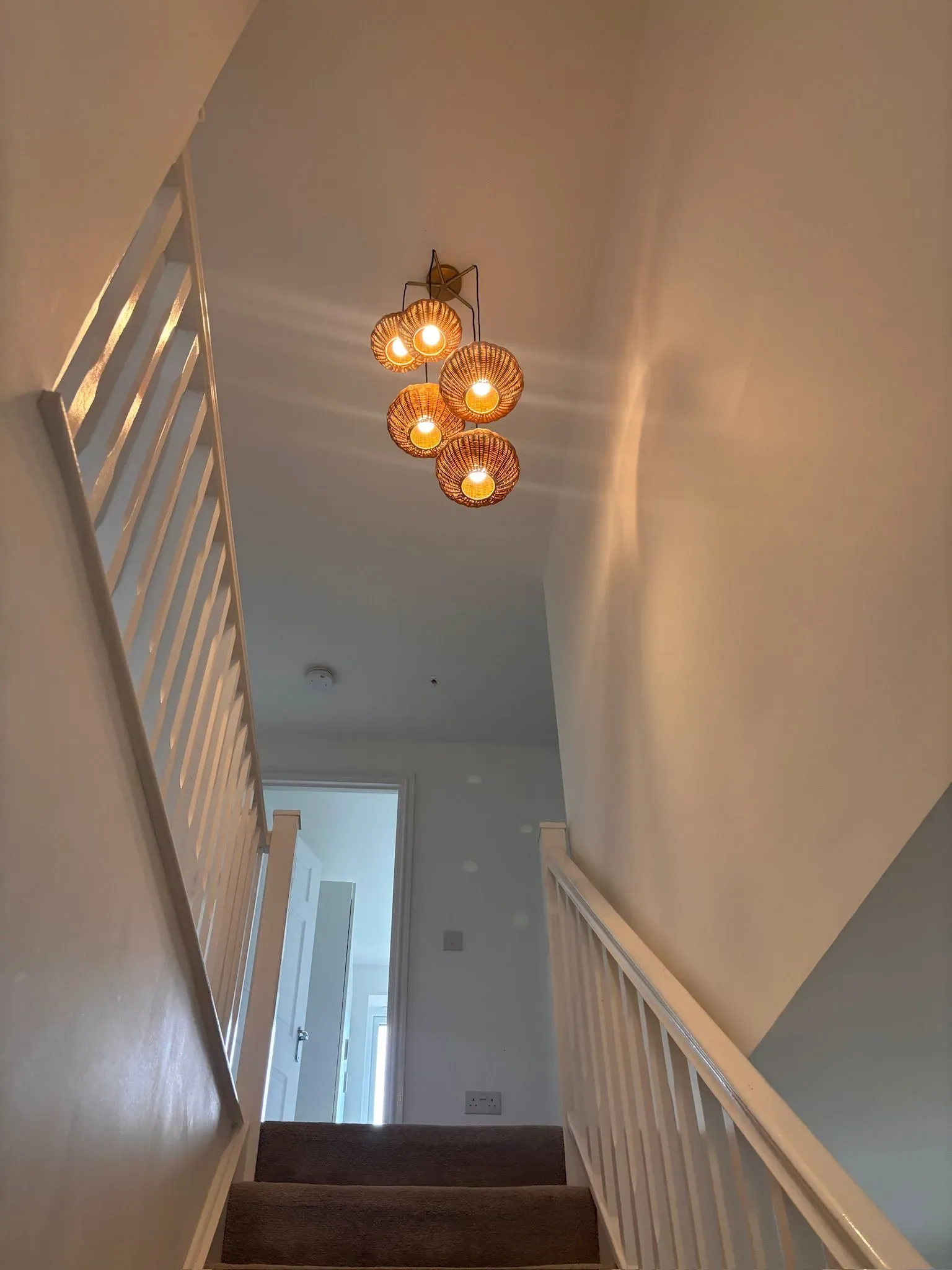 A cluster of five woven light pendants hanging from the ceiling in a hallway above a staircase with white railings.